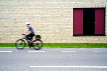 Person riding a bicycle past a brick wall with a large red window, motion blur conveys movement and...