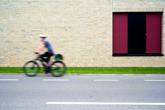 Person riding a bicycle past a brick wall with a large red window, motion blur conveys movement and speed, adding dynamism to the scene.