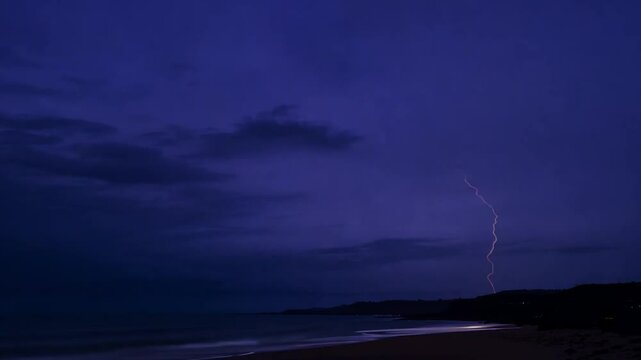 A vibrant display of lightning arcs across a darkening sky as it nears evening. The tranquil ocean reflects the stormy atmosphere, blending with the gentle waves on the shore.