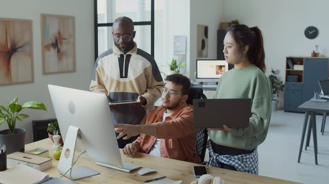 Medium full shot of three diverse male and female programmers, holding tablet, laptop, standing by colleagues desk, looking at computer screen, discussing work on AI project code, in startup office