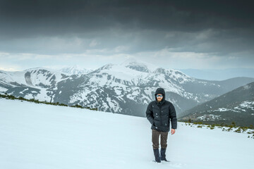 Stylish young man with a beard hiking in snowy mountain area wearing cold weather accessories, warm down jacket against a scenic backdrop of snowy mountains and  snow falling from overcast skies.