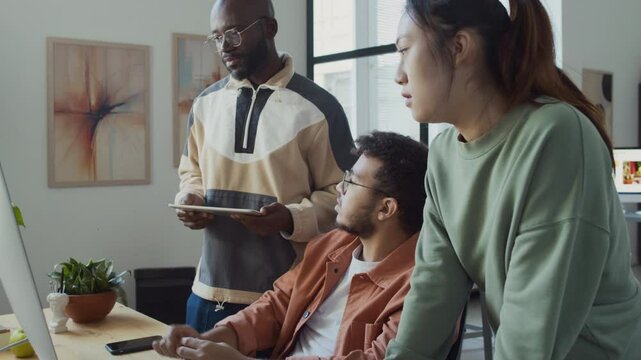 Medium pan shot of young Middle Eastern male software developer suggesting solution to bug in code to Chinese female and black male colleagues, while discussing AI image generation project in office