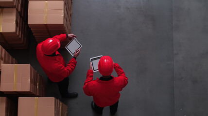 Employees in red jackets and helmets using tablets to manage inventory in warehouse filled with stacked boxes, showcasing modern logistics and teamwork