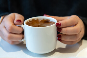 Close-up view of female hands with red fingernails holding white blank porcelain cup of americano coffee with brown crema by white table. Soft focus. Copy space. Hot drinks business theme.