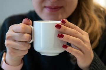 Close-up view of female hands with red fingernails holding white blank porcelain cup of coffee or tea by her face. Soft focus. Copy space. Hot drinks business theme.