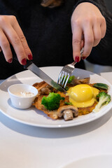 Close-up view of woman eating healthy breakfast: boiled egg, fried yam with marrow pancake, fried thin croissant, broccoli, mushrooms, yellow hollandaise sauce and white sour cream (smetana).