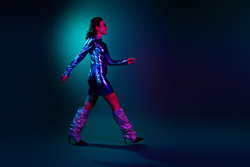 Full size profile photo of gorgeous young girl walk empty space wear shiny dress neon light dark blue background