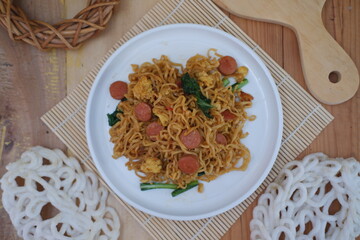 A plate of seasoned instant noodles is garnished with sliced sausage, leafy greens, and a piece of crispy cracker.