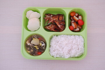 A green tray with five compartments containing white rice, curry, sliced cucumbers and tomatoes, a boiled egg, quail eggs, fried chicken, and pieces of meat