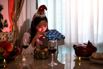Asian young woman in festive headband looks into glowing gift box at Christmas party dinner table with decorations and food