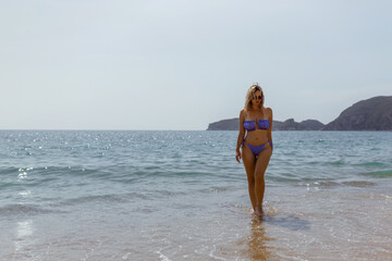 amazing girl with beautiful athletic body walking and posing on white sand beach. Tanned young woman with curly hair relaxes and sunbathes. Charming model posing nearby