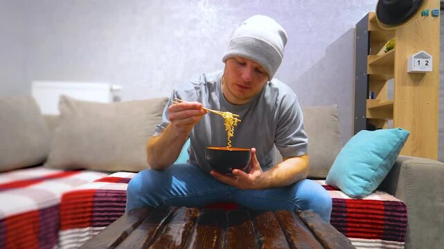A young man eats doshirak noodles with wooden chopsticks. Eating pasta at home at the table in the living room. Slow motion.