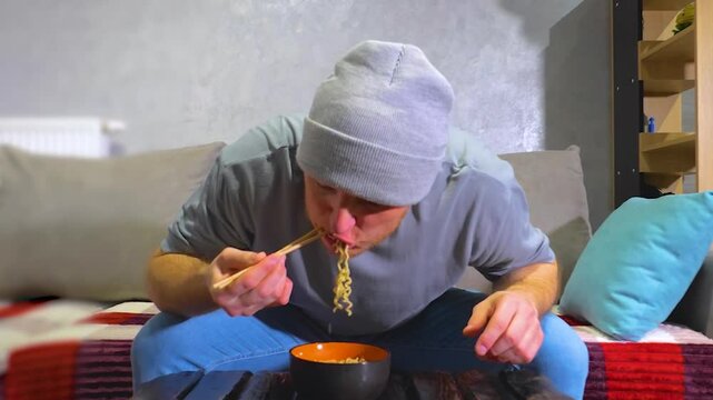 Stylish young man eats doshirak noodles with wooden chopsticks. Eating pasta at home at the table in the living room. Slow motion