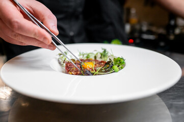 A chef garnishes a gourmet dish with tweezers, focusing on the intricate details of plating. The scene captures the artistry and precision of high-end culinary presentation in a professional kitchen.