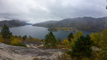 Scenic view of a fjord surrounded by rocky cliffs and autumn trees under a cloudy sky.