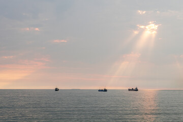 Tranquil ocean at sunset with sun rays breaking through clouds, lighting up three distant boats on calm waters. A peaceful seascape with soft light, creating a serene maritime view.