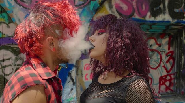 Gothic girl puffs cigarette smoke in the face of a redhead punk-style guy. A close-up of a couple sharing a smoke kiss in an urban, graffiti-covered setting.
