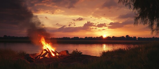 Fototapeta premium Cozy bonfire by the river during a vibrant summer sunset with dramatic clouds reflecting on the water surface.