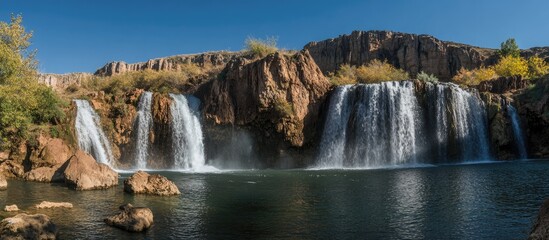 Fototapeta premium Serene waterfalls cascading into a tranquil pool surrounded by rocky cliffs and lush greenery under a clear blue sky