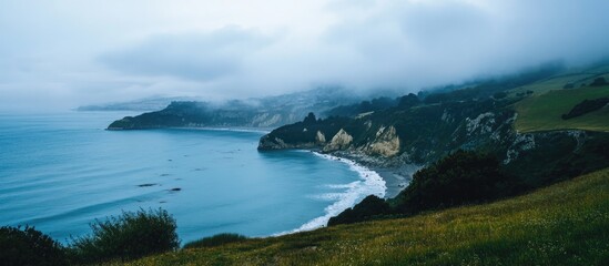 Fototapeta premium Serene coastal landscape with misty sea and rolling hills under a cloudy sky showcasing nature's tranquility and beauty