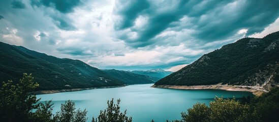 Serene panorama of a mountain lake surrounded by lush hills under a dramatic cloudy sky with turquoise waters reflecting the landscape.