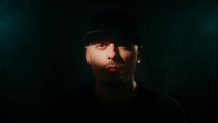 A man in wearing a black hat, lit with moody and dramatic lighting against a dark background.