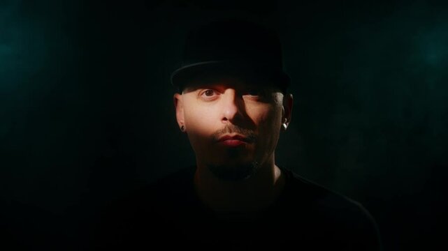A man in wearing a black hat, lit with moody and dramatic lighting against a dark background.