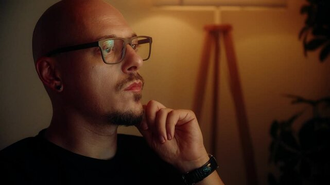 A man wearing glasses and a black shirt sitting in fron of monitor in a warmly lit room with a lamp and indoor plants in the background.