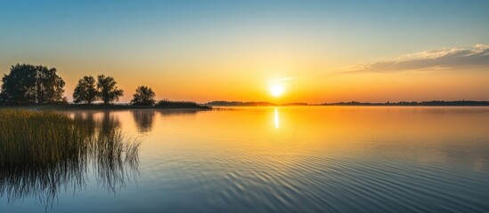 Obraz premium Serene sunrise over tranquil waters with silhouetted trees reflecting warm hues of orange and blue in a peaceful landscape.
