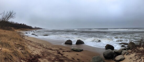 Obraz premium Serene winter beach scene with gray skies and turbulent waves crashing against rocky shoreline and sandy beach.
