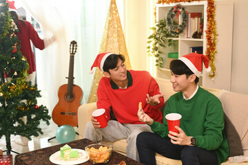Two friends in festive hats chat cheerfully on a cozy couch, having conversations and enjoying snacks and drinks at the Christmas party at home