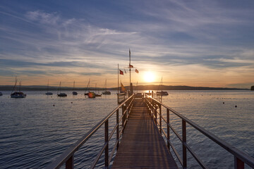 Obraz premium A serene sunset over a calm river, with vibrant orange and pink hues in the sky. A small sailboat is anchored in the water, reflecting the colorful sky.