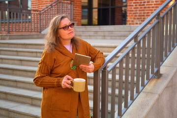 Middle-aged woman in glasses holding a tablet and cup of coffee while standing on stairs. Outdoor...