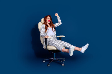 Fototapeta premium Young woman happily reclining in an office chair against a dark blue background, wearing a trendy checkered suit and white sneakers