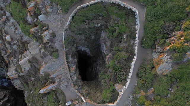 Aerial view of Ons Island's Burato do Inferno, a deep cavern descending to the sea, bordered by stone wall and surrounded by lush vegetation. The Atlantic waters are visible at the end of the cavern.