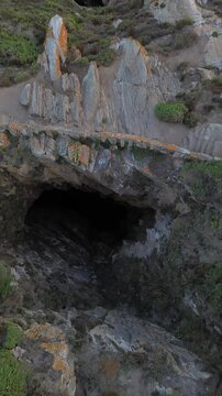 Aerial view of Ons Island's Burato do Inferno, a deep cavern descending to the sea, bordered by stone wall and surrounded by lush vegetation. The Atlantic waters are visible at the end of the cavern.