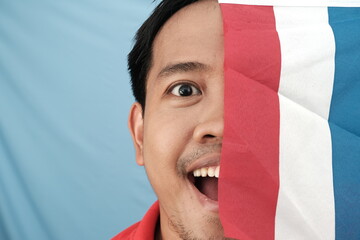 Close-up of a cheerful man peeking behind a Dutch flag, expressing excitement and playfulness...