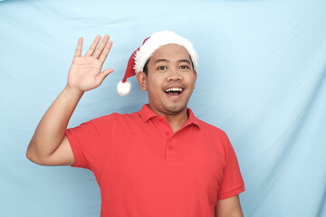 Cheerful Asian man in a red shirt and Santa hat waving with an excited expression, standing against a light blue background