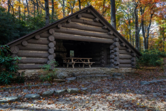 A rustic log cabin sits in a forest, surrounded by colorful autumn leaves. Sunlight filters through the trees, highlighting the picnic table inside the open structure. - Powered by Adobe