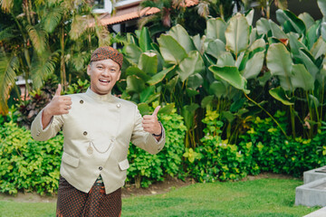 Joyful Asian man wearing traditional Javanese beskap and batik is giving a thumb up gesture.