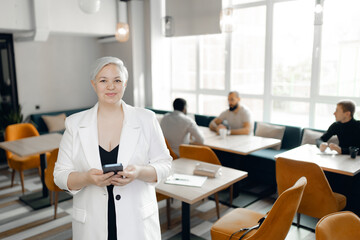 Short-haired caucasian woman in white blazer holding smartphone in contemporary cafe surrounded by colleagues. bright natural lighting emphasizes collaborative and professional atmosphere.