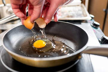 A pair of hands cracking an egg over a frying pan, with the yolk and some egg white already in the...