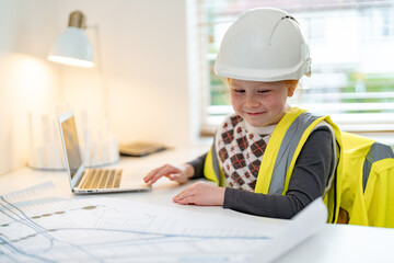 Young child in construction gear working on plans with laptop at a desk indoors