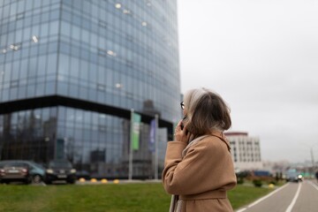 A woman in a beige coat talks on her phone while standing near a modern glass office building on a...