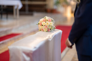 A delicate bridal bouquet with white and pink roses placed on a satin-covered church bench, with a...
