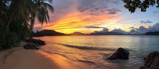 Breathtaking beach sunset over calm waters with palm trees and rocky shoreline creating a serene tropical landscape at dusk.