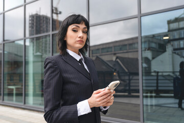 Confident businesswoman standing outdoors with smartphone in hand near urban glass building