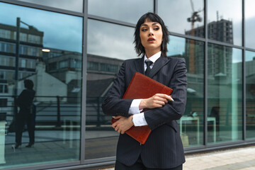 Fototapeta premium Confident professional woman in suit holds folder outside modern office building during cloudy day