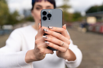 Person takes a selfie in an outdoor setting with a smartphone and stylish nails on a cloudy day