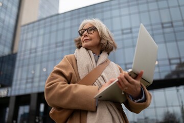 Senior woman using laptop outdoors in modern urban setting on a cloudy day while enjoying her work and the fresh air